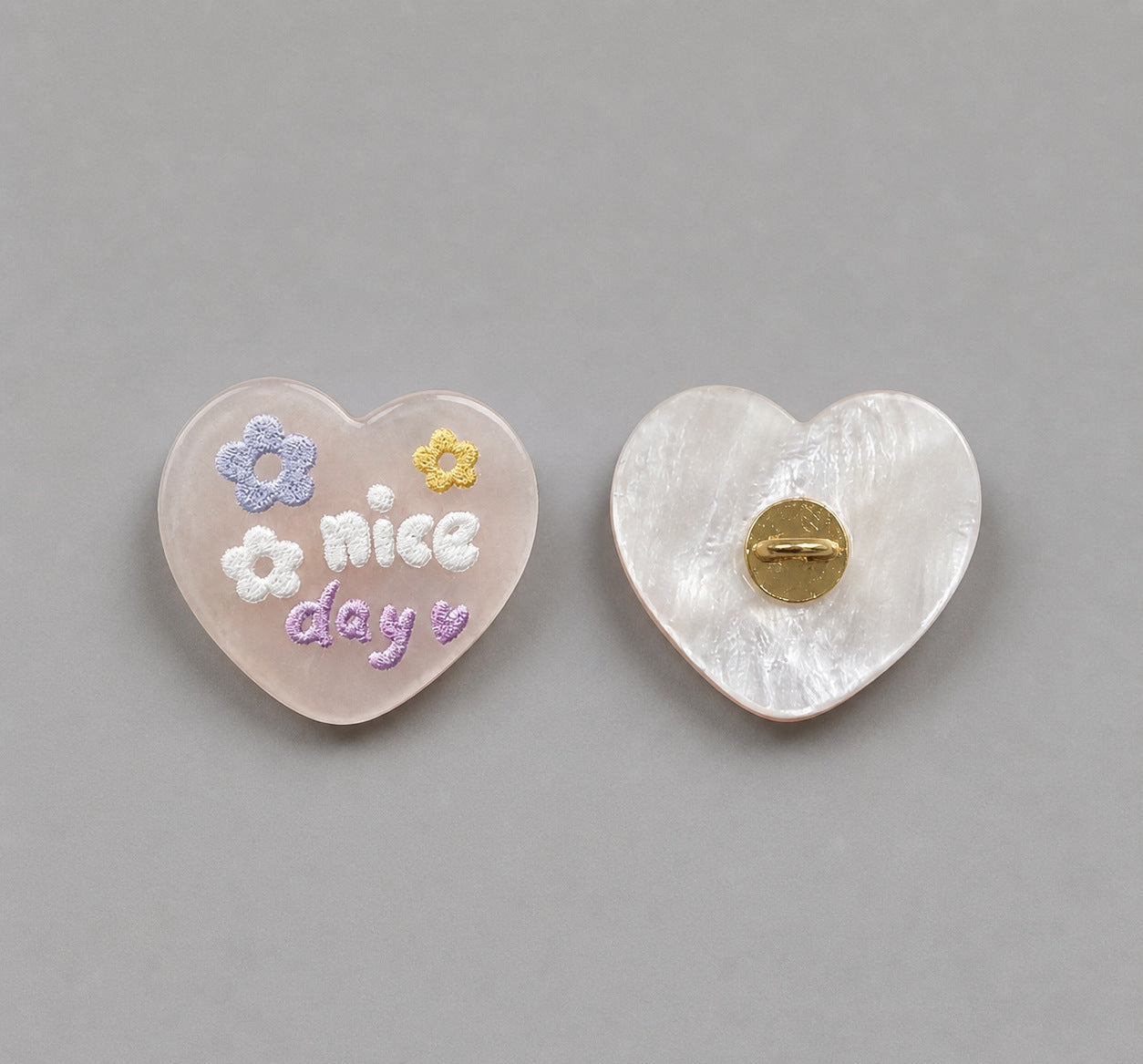 Light beige heart-shaped resin button with “nice day” and flowers; white back with gold pin