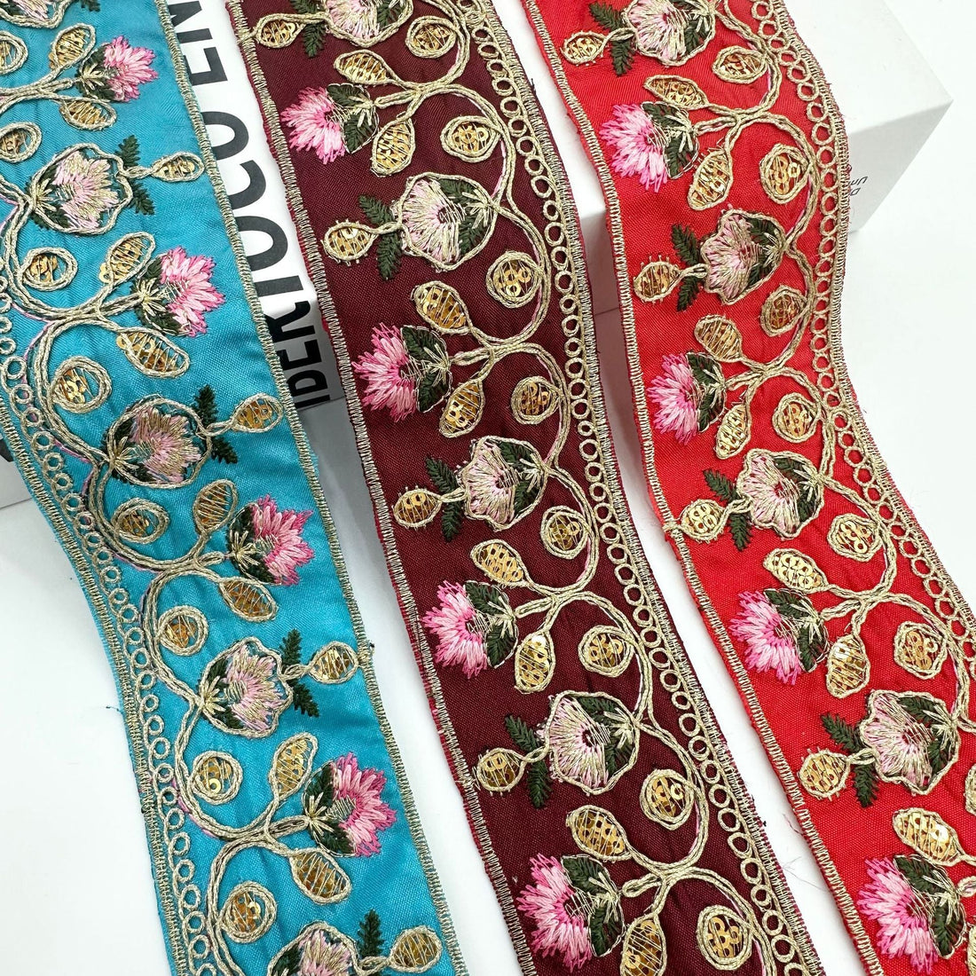 Embroidered lace trim ribbon with pink flowers, gold motifs, and teal fabric background